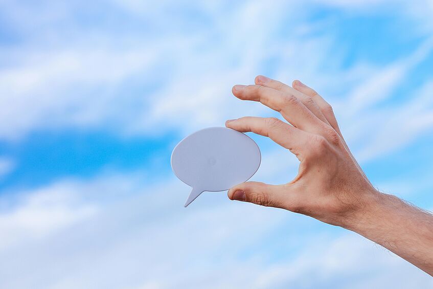 A hand holding a paper in the shape of a speech bubble against the sky.